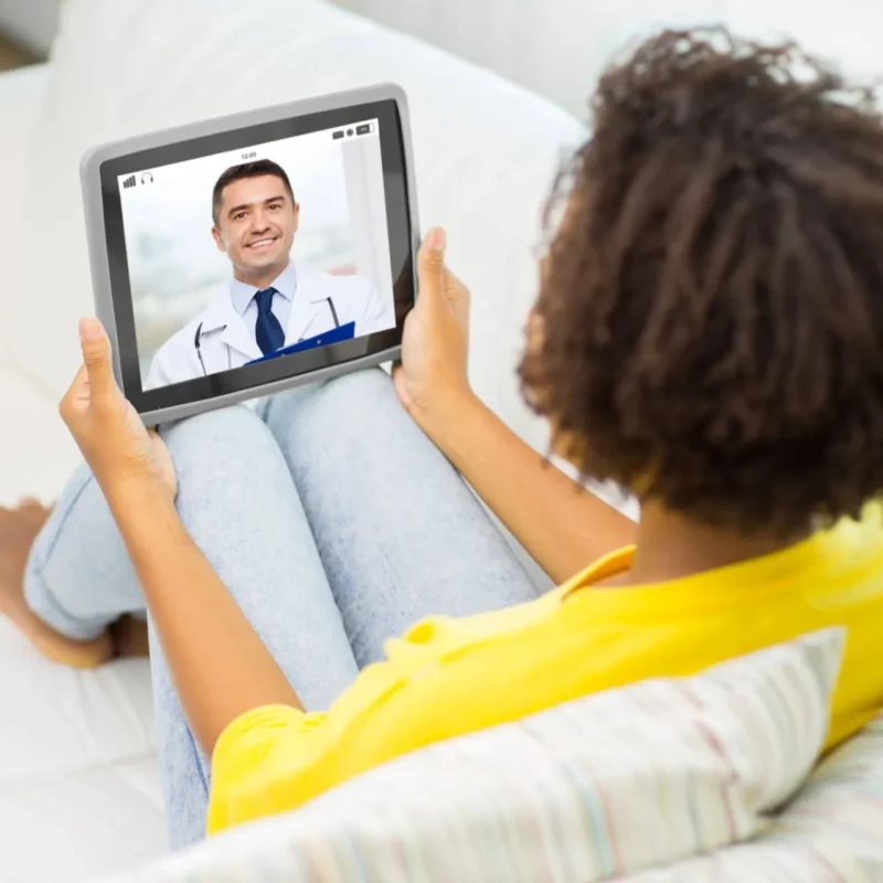 acidente pessoal: Paciente deita em uma cama e fala por videochamada em tablet com um médico, enquanto recebe atendimento à distância em ambiente doméstico.