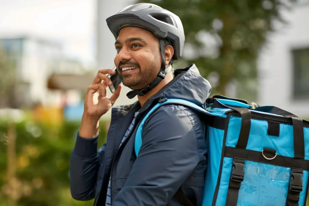 svg+xml;charset=utf em Maximídia - Seguro de Acidentes Pessoais e Seguro de Vida com o Máximo de Benefícios Homem com capacete e mochila azul falando ao celular ao ar livre, em ambiente urbano, sugerindo contratação de seguro acidente durante atividades diárias