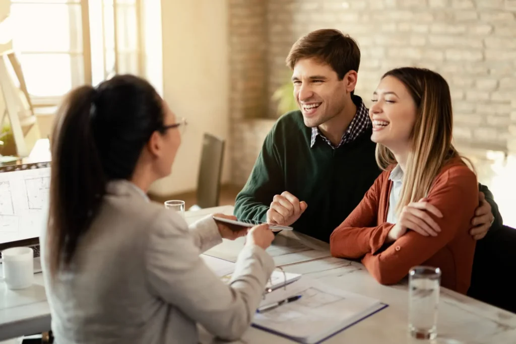 svg+xml;charset=utf em Maximídia - Seguro de Acidentes Pessoais e Seguro de Vida com o Máximo de Benefícios Corpo de consultor e casal sorridente em reunião à mesa com contrato e tablet, discutindo seguro e proteção financeira em ambiente residencial. seguro de vida e acidentes pessoais